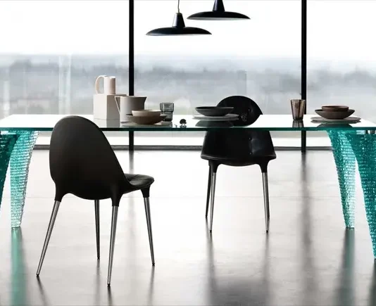 Mesa Atlas de vidrio con patas esculpidas, rodeada de sillas negras en un comedor moderno.
