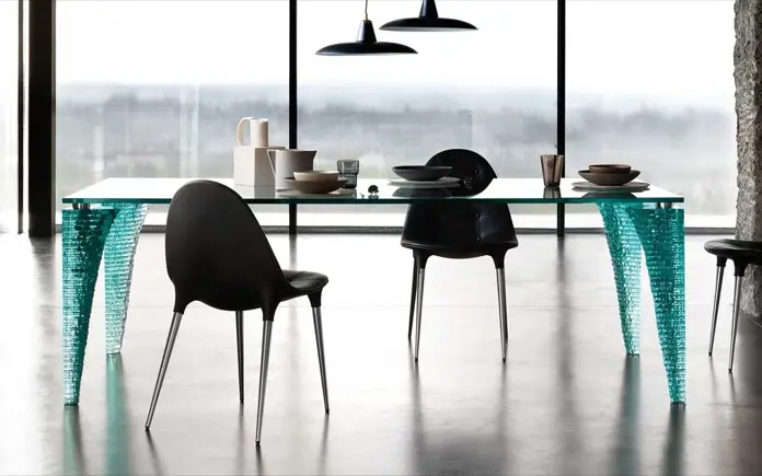 Mesa Atlas de vidrio con patas esculpidas, rodeada de sillas negras en un comedor moderno.